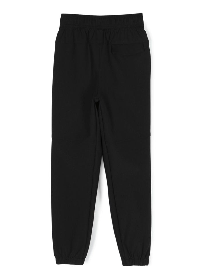 Pantalon de sport noir et blanc pour enfant, détail plissé, logo imprimé sur la jambe, taille haute, fermeture sur le devant avec boutons et zip, deux poches latérales en biais et poignets élastiqués